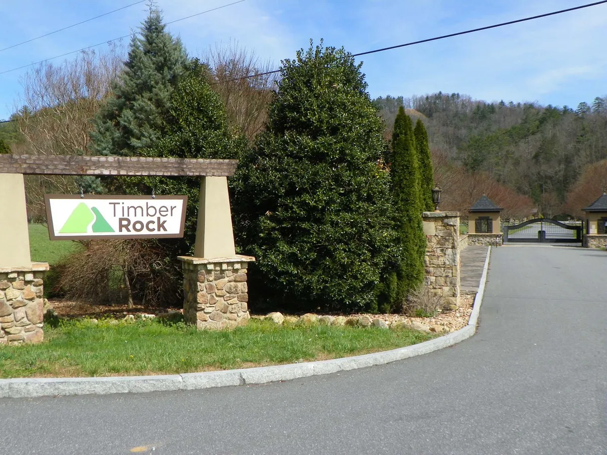 The gated entrance to the community, featuring the Timber Rock sign set among landscaped greenery. The road beyond the gate hints at a secluded, secure setting tucked into natural surroundings.