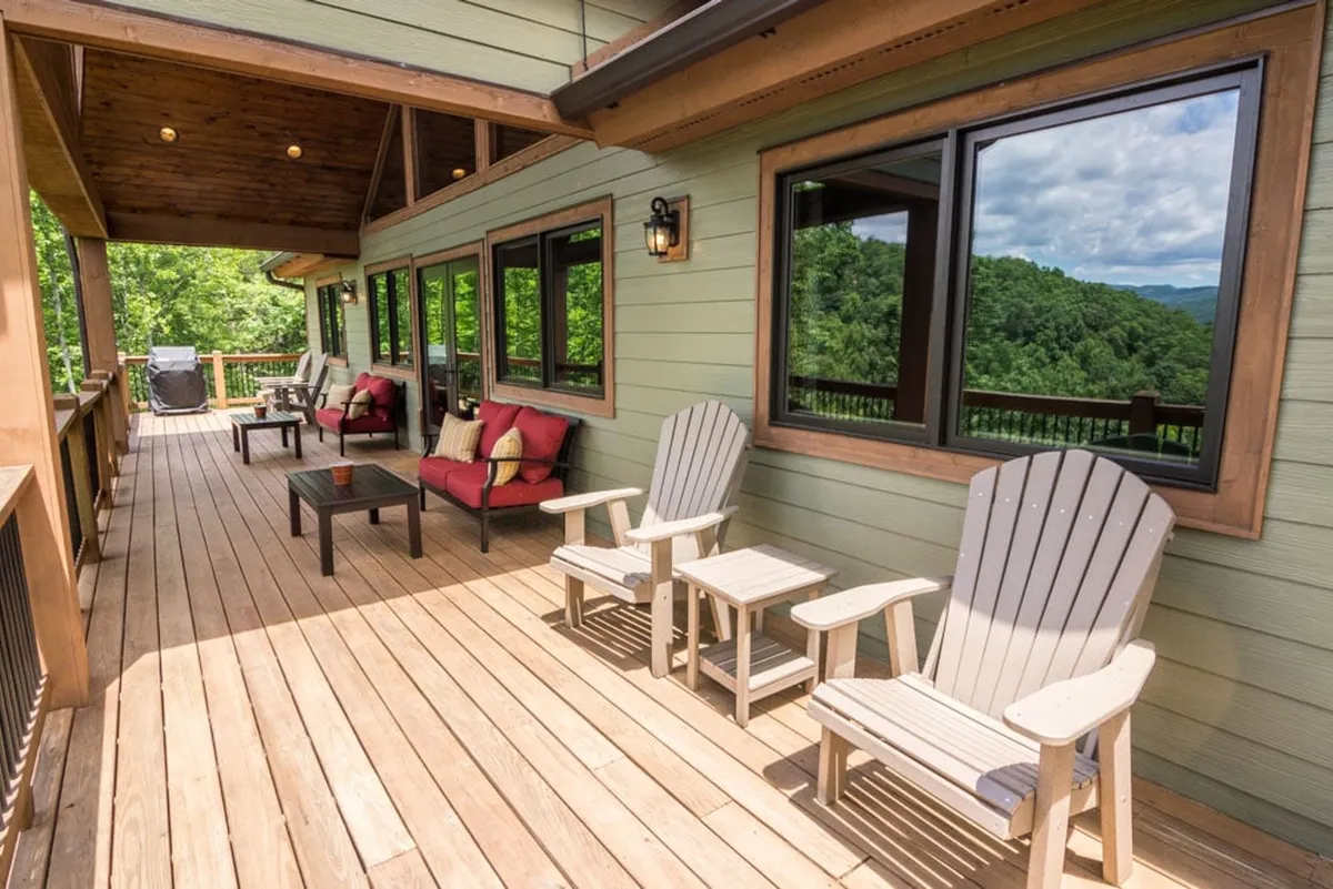 A spacious wooden deck with comfortable seating overlooks lush green landscapes and distant mountains, with large windows adding a sense of connectivity between the home and nature. The setting is ideal for relaxing after outdoor activities, sharing laughter with friends, or simply soaking in the tranquil environment.