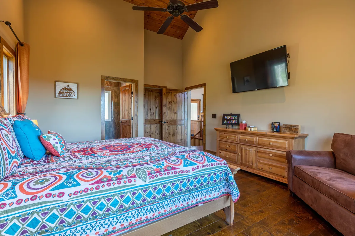 A bright, inviting bedroom with a colorful bedspread, wooden dresser, TV on the wall, and windows allowing natural light to stream in. The room feels serene and comfortable, ideal for both rest and relaxation after a day of adventure.