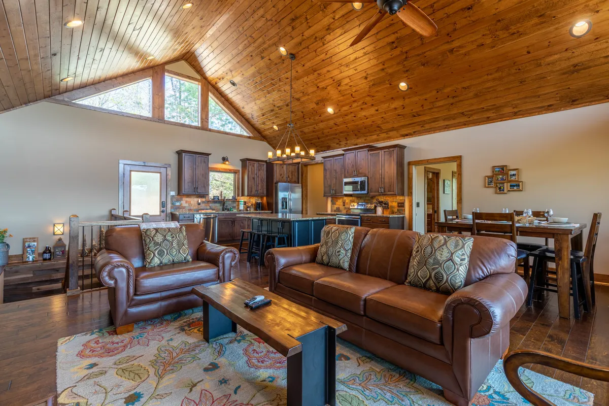 A cozy, inviting angle on the great room reveals rich leather sofas, a warm wooden ceiling with timber beams, and a view into the open kitchen with a substantial island. The arrangement encourages relaxation and conversation, highlighting both comfort and communal gathering.