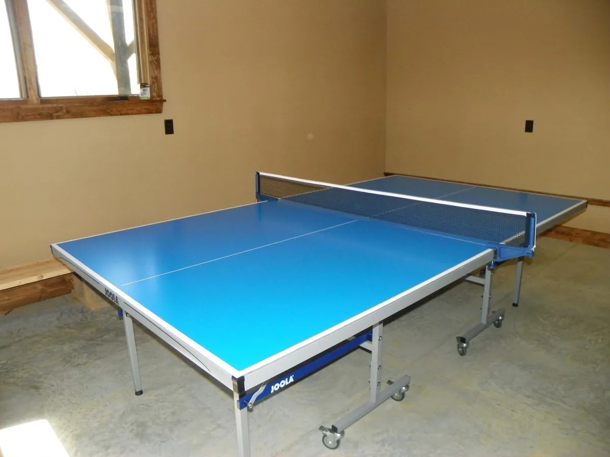 This image features a garage space set up with ping pong tables under exposed wooden beams, illuminated by natural light streaming through a window. The scene exudes an atmosphere of playfulness and casual fun, ideal for friendly tournaments or game nights.