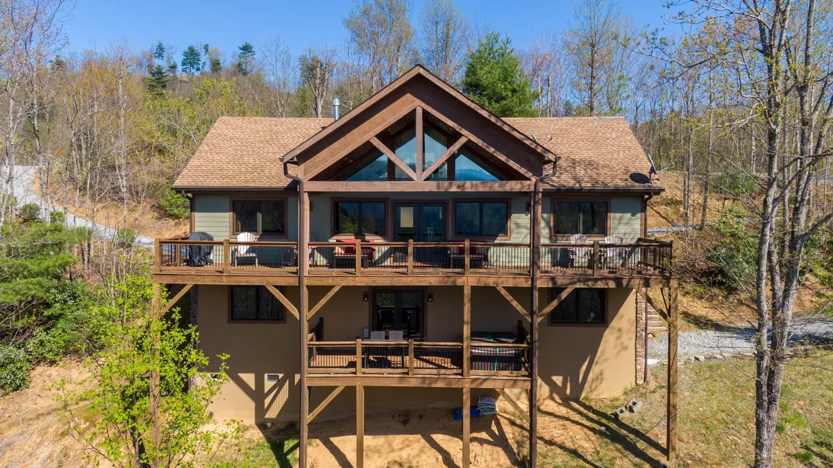 The image showcases the exterior of a stunning Timberframe-style house with a striking triangular roof and expansive windows, all set against a backdrop of breathtaking mountain views. The spacious deck invites relaxation and entertaining, with sunset colors spilling over the horizon and the promise of starlit evenings.