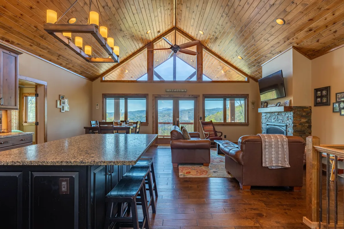 A bright, expansive great room featuring timberframe construction, large picture windows with sweeping mountain views, a cozy seating area with plush sofas positioned near a stone fireplace, and a spacious kitchen island with barstools set for casual dining. The open layout seamlessly connects the living, dining, and kitchen spaces.