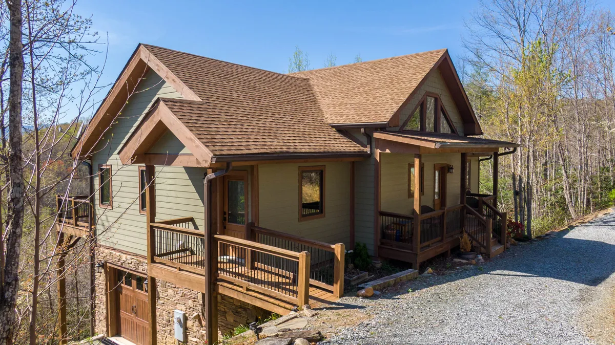 The image highlights the home's facade and spacious deck surrounded by lush greenery. The inviting deck offers plenty of room for family gatherings, barbecues, and evenings spent stargazing or soaking in the mountain air.