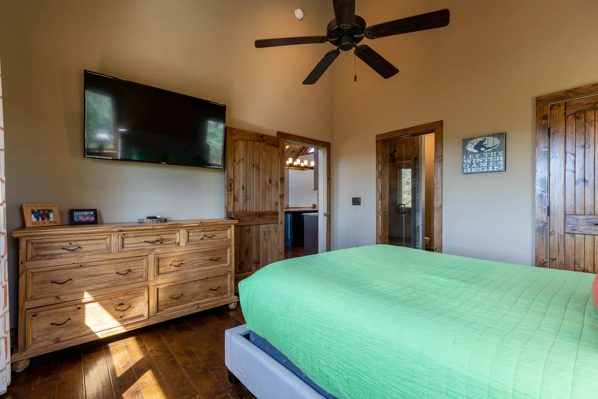 A restful bedroom with a green bedspread, wooden dresser, and a wall-mounted TV. The space is styled for comfort, encouraging relaxing, watching TV, or reading. The overall look is modern yet cozy, appealing to anyone seeking a private place to recharge.