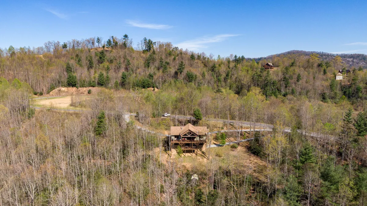 This image showcases the wooden house nestled among tall trees with a winding road leading up to it. The tranquil exterior scene hints at a secluded mountain retreat, perfect for family gatherings and outdoor activities such as stargazing and morning coffee with mountain views.