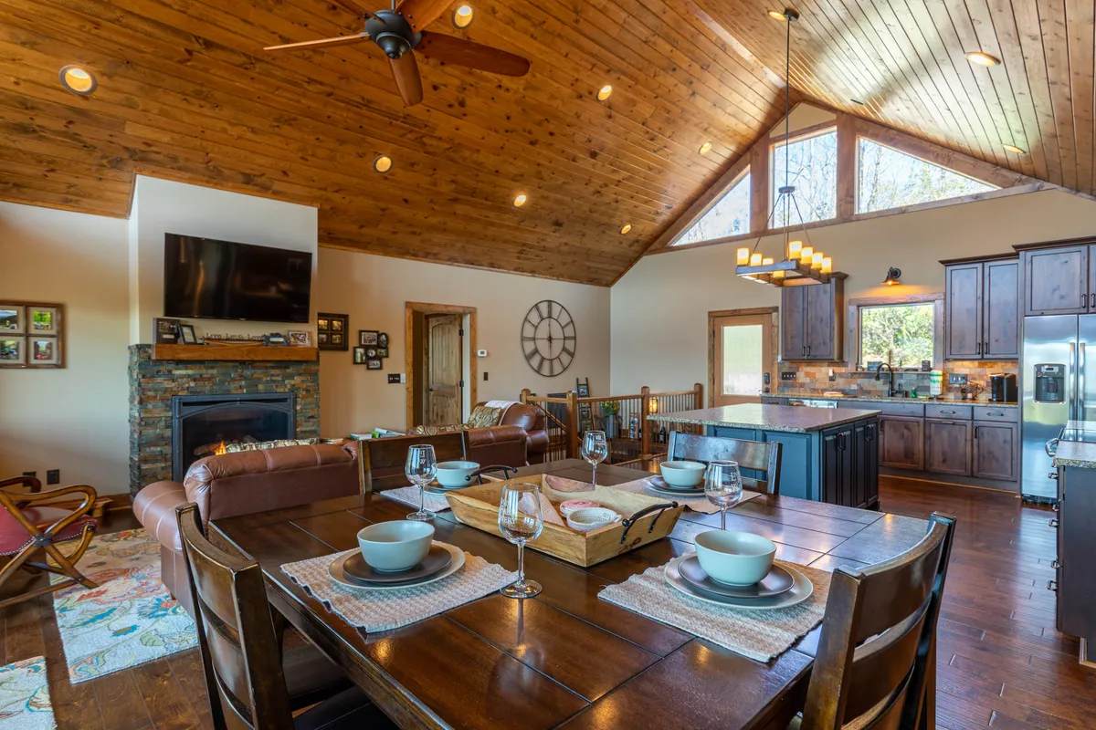 The dining area is elegantly set for a meal, with a large dining table, stone fireplace, expansive windows letting in natural light, and a glimpse of the modern kitchen. The scene radiates a sense of welcoming hospitality and is ideal for family dinners or festive celebrations.